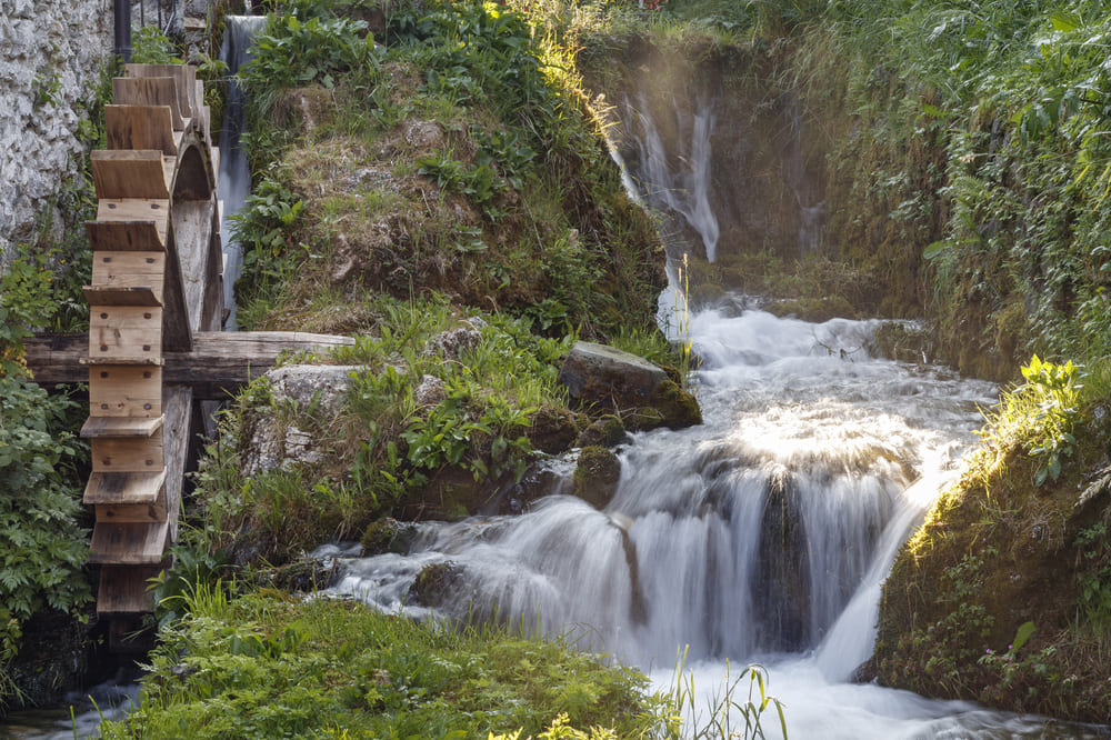 Antico mulino ad acqua in pietra con grande ruota in legno, situato lungo il rio Touf a Illegio, immerso in un paesaggio verde e tranquillo.