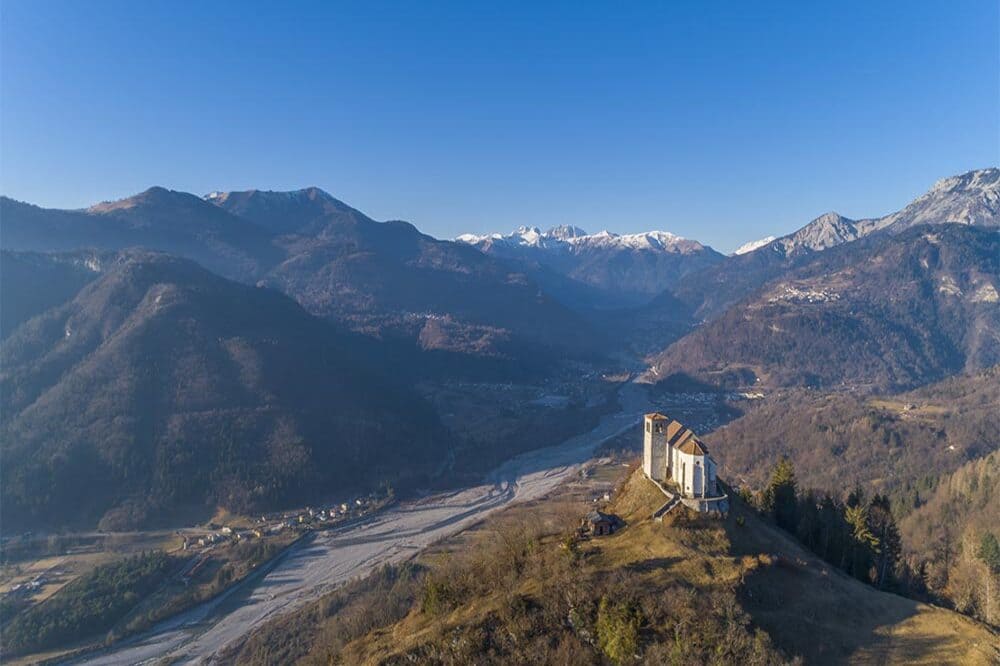 La Pieve di San Floriano in cima al monte Gjaideit, a Illegio, con vista panoramica sulle montagne, circondata da una natura rigogliosa.