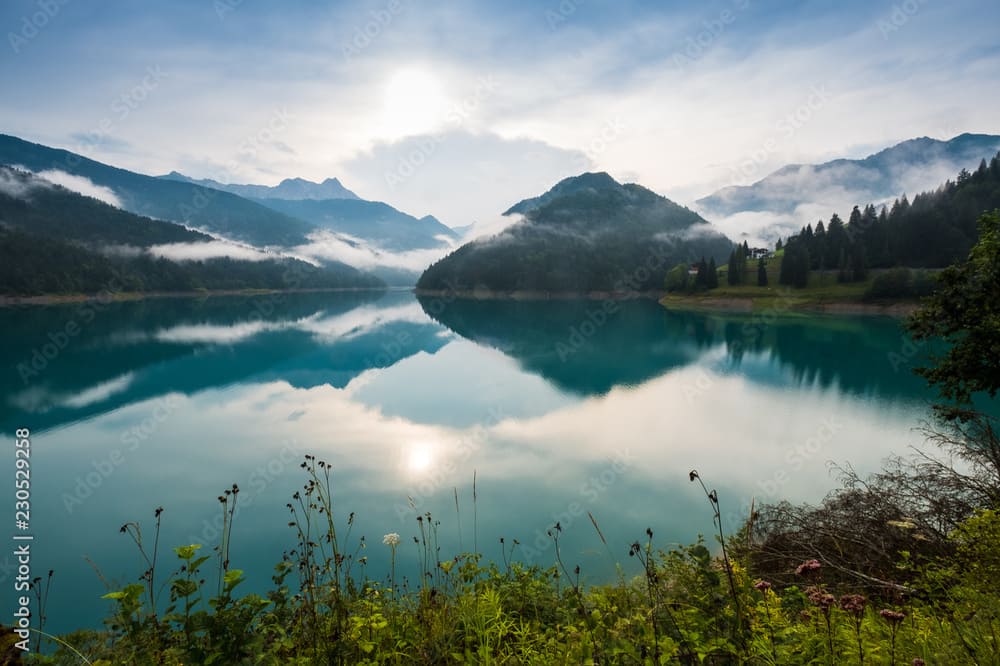 Lago di Sauris all'alba con nebbia e montagne riflettenti nell'acqua